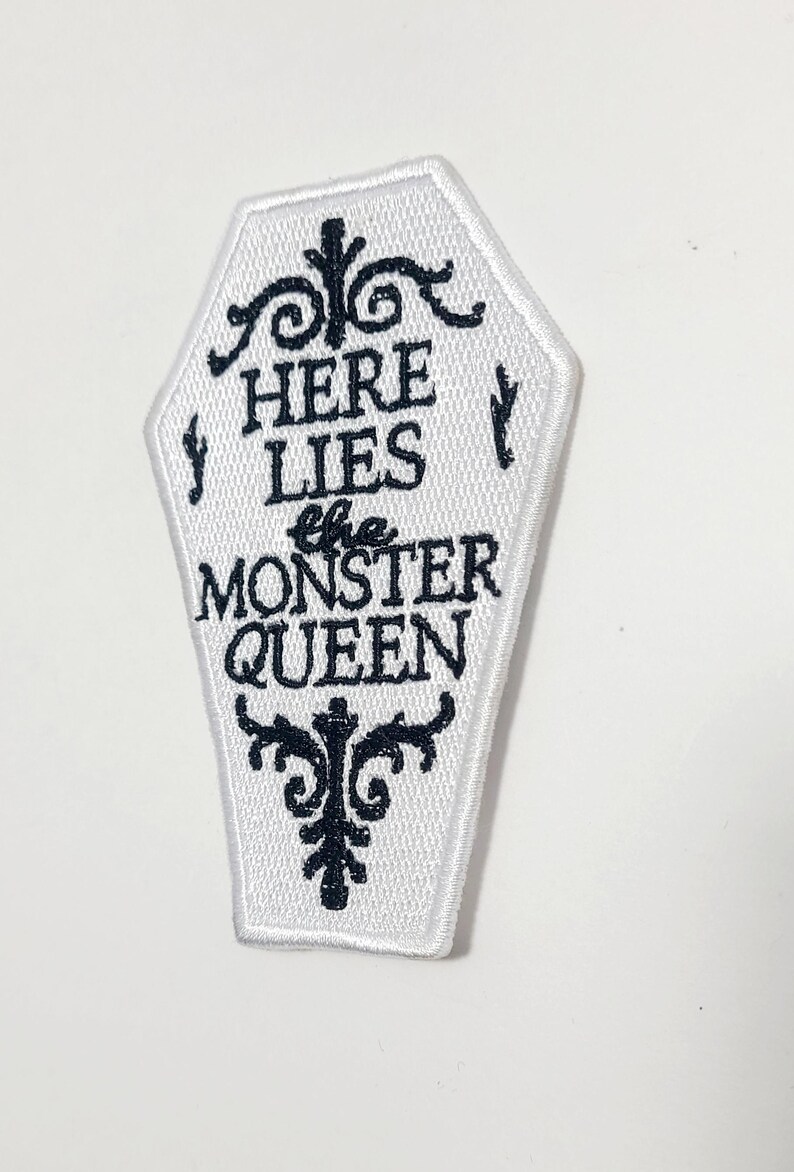 Puede incluir: Parche blanco en forma de ata&uacute;d con texto bordado en negro y adornos decorativos. El texto dice "HERE LIES the MONSTER QUEEN". Un parche de tem&aacute;tica g&oacute;tica y espeluznante para ropa o accesorios.