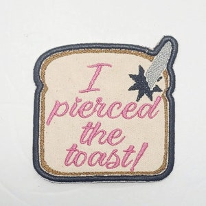 Può includere: Una toppa ricamata a forma di fetta di pane, con un contorno nero e un bordo dorato. La toppa è beige e presenta il testo "I pierced the toast!" in corsivo rosa. Un coltello argentato con una stella nera è conficcato nel pane.