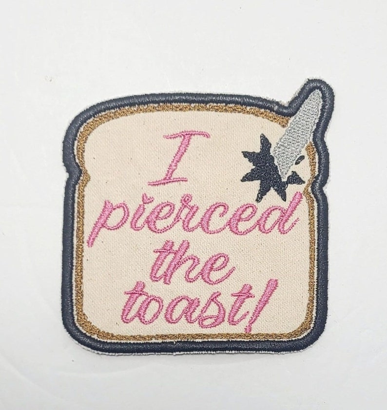 May include: A beige embroidered patch with a black outline and gold trim. The patch is shaped like a slice of bread and features the text "I pierced the toast!" in pink cursive. A silver knife with a black star is sticking into the bread.