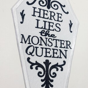 Puede incluir: Parche blanco en forma de ata&uacute;d con texto bordado en negro y adornos decorativos. El texto dice "HERE LIES the MONSTER QUEEN". El parche tiene un borde blanco, para un tema de Halloween o g&oacute;tico.