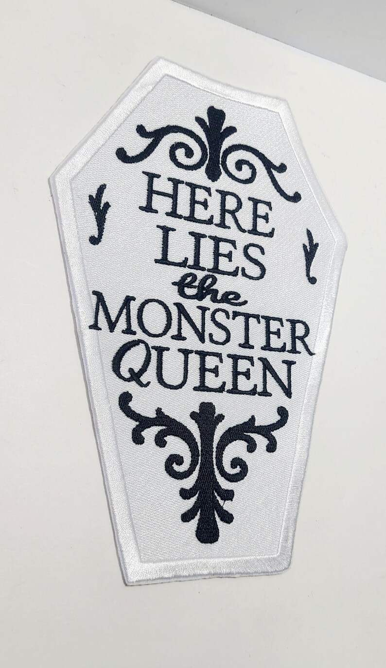 Puede incluir: Parche blanco en forma de ata&uacute;d con texto bordado en negro y adornos decorativos. El texto dice "HERE LIES the MONSTER QUEEN". El parche tiene un borde blanco, para un tema de Halloween o g&oacute;tico.