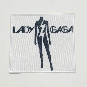 Puede incluir: Un parche cuadrado blanco con las palabras "LADY GAGA" en negro, con una figura negra estilizada y un diseño de rayo. El parche tiene una textura tejida y un diseño limpio.
