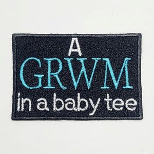 May include: A rectangular patch with the text "A GRWM in a baby tee." The patch has a black background with white and teal lettering. The patch has a black border.