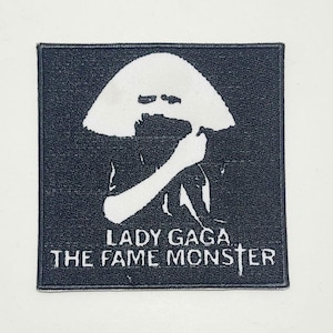 Könnte beinhalten: Schwarzer quadratischer Aufnäher mit einer weißen Silhouette einer Person mit großem Hut. Der Text "LADY GAGA THE FAME MONSTER" ist darunter in Weiß gedruckt. Für Kleidung geeignet.