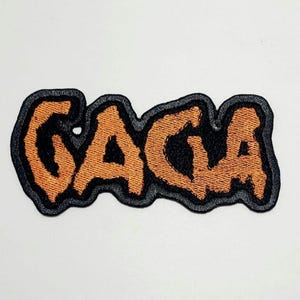 May include: Embroidered patch featuring the word "GAGA" in orange, with a black outline and a dark grey border. The patch has a textured finish and is set against a white backdrop.