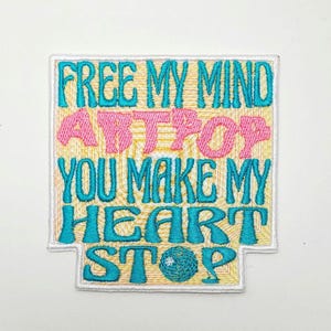 May include: Embroidered patch with the words "FREE MY MIND ARTPOP YOU MAKE MY HEART STOP". The text is in teal and pink on a yellow background, with a white border. A small teal and white design is at the bottom.