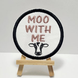 May include: A white round patch with black edging, featuring the text "MOO WITH ME" in pink embroidery and a black embroidered cow head.