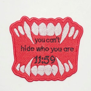 Op de afbeelding: Rode geborduurde patch met een set witte tanden en de tekst "you can't hide who you are 11:59".
