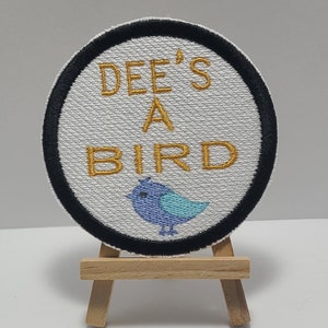 May include: A white round patch with a black border, featuring the text "DEE'S A BIRD" in gold embroidery. A blue and turquoise embroidered bird is centered below the text.