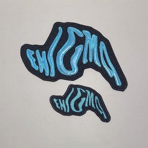 Può includere: Due toppe nere ricamate con la parola "ENIGMA" in un carattere stilizzato, blu chiaro e lucido. Le toppe hanno una forma unica, una più grande dell'altra. Lo sfondo è grigio chiaro.