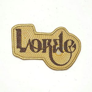 May include: Embroidered patch in a key-shaped design. The patch features the word "Lorde" in a dark brown, stylized font, set against a gold-colored background. The border is also gold, creating a textured appearance.