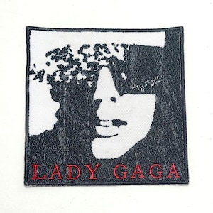 May include: Square black and white patch featuring a stylized portrait of Lady Gaga. The image is a black and white design with the text "LADY GAGA" embroidered in red at the bottom. The patch has a black border.