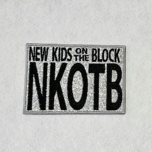 May include: A rectangular embroidered patch featuring a silver glitter background with black stitched lettering. The text reads NEW KIDS ON THE BLOCK in a smaller font above the large, bold acronym NKOTB, all enclosed within a thin black border.