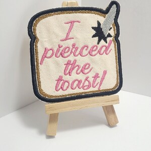 May include: A white embroidered patch with black and gold trim, shaped like a slice of bread. The patch says "I pierced the toast!" in pink thread. The patch is displayed on a small wooden easel.