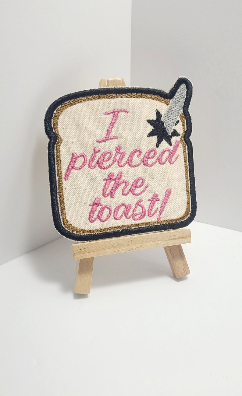 May include: A white embroidered patch with black and gold trim, shaped like a slice of bread. The patch says "I pierced the toast!" in pink thread. The patch is displayed on a small wooden easel.