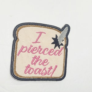 May include: A beige embroidered patch shaped like a slice of bread with the text "I pierced the toast!" in pink thread. A silver knife is sticking out of the top of the bread.