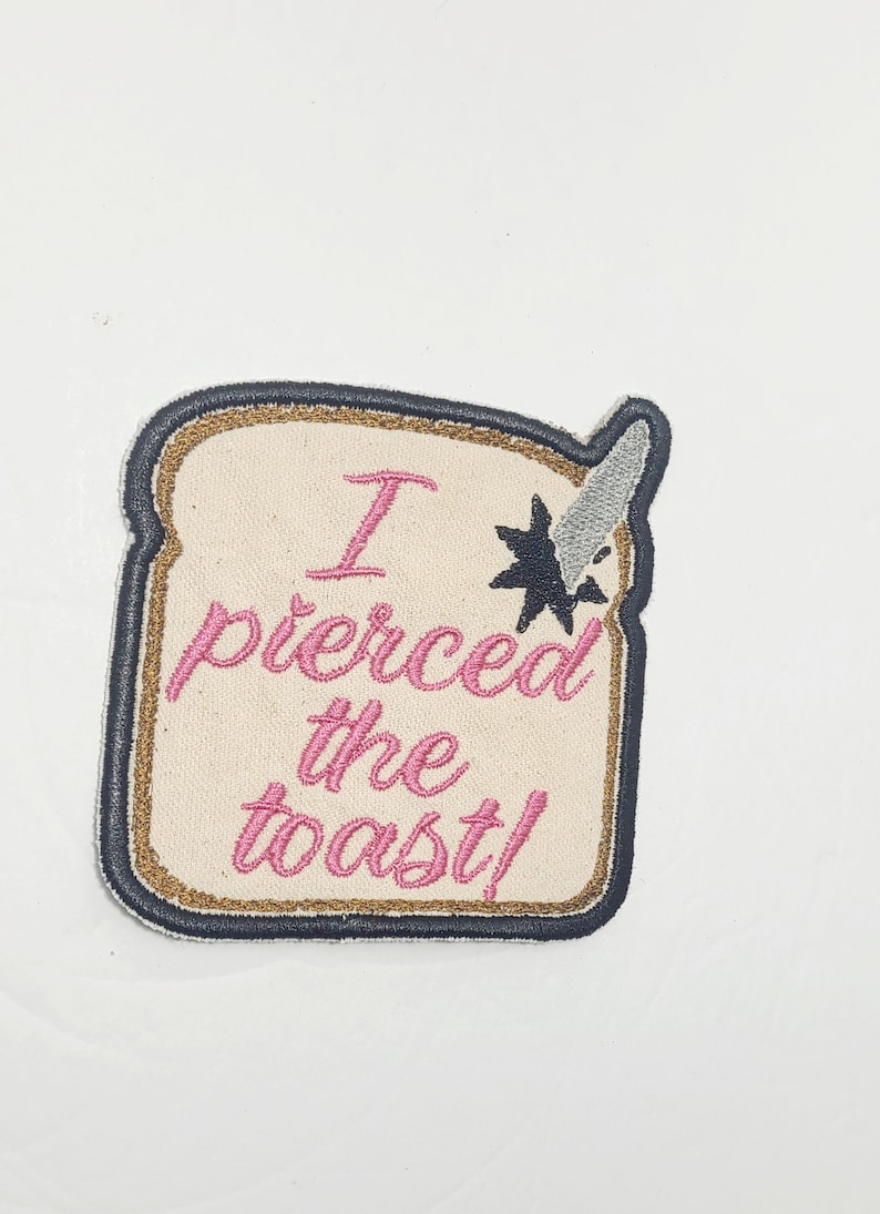 May include: A beige embroidered patch shaped like a slice of bread with the text "I pierced the toast!" in pink thread. A silver knife is sticking out of the top of the bread.