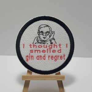 May include: A white embroidered patch with a black border. The patch features a cartoon drawing of a person wearing glasses and the text "I thought I smelled gin and regret".