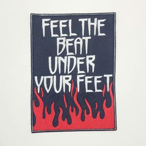 May include: A rectangular patch with the text "FEEL THE BEAT UNDER YOUR FEET" in white, embroidered on a navy blue background. The bottom edge features a red flame design.