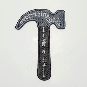 May include: Embroidered patch in the shape of a hammer. The hammer head has the words "everything looks" in white thread. The handle has the words "like a nail" in white thread. The patch is black.