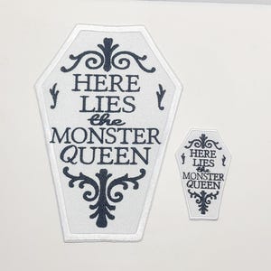 May include: Two white coffin-shaped patches with black text and decorative flourishes. The larger patch reads "HERE LIES the MONSTER QUEEN." The smaller patch mirrors the design. The patches are likely for clothing or accessories.