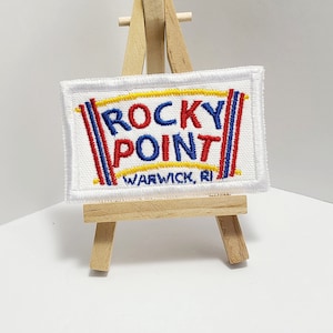 May include: White embroidered patch with the text "ROCKY POINT" in red, blue, and yellow lettering. The text "WARWICK, RI" is embroidered in black below the main text.