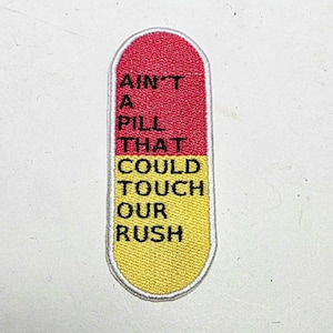 Könnte beinhalten: Ein gestickter Aufnäher in Pillenform, unterteilt in rote und gelbe Abschnitte. Der Aufnäher trägt den Text "AIN'T A PILL THAT COULD TOUCH OUR RUSH" in Schwarz. Er hat einen weißen Rand.