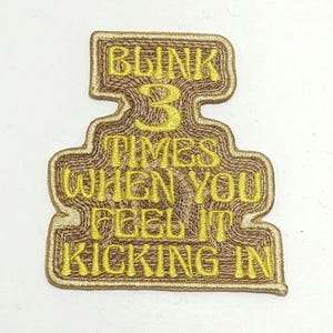 Könnte beinhalten: Gestickter Aufnäher mit dem Text "BLINK 3 TIMES WHEN YOU FEEL IT KICKING IN" in gelbem Faden auf braunem Hintergrund. Der Aufnäher hat eine einzigartige, geschichtete Form mit strukturiertem Aussehen, geeignet für Kleidung oder Accessoires.
