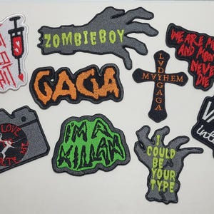 Puede incluir: Una colecci&oacute;n de parches bordados con varios dise&ntilde;os y texto. Los parches incluyen frases como "I just need a hit", "Zombieboy" y "Gaga". Otros parches presentan una c&aacute;mara, una cruz y una mano con texto. La paleta de colores incluye negro, gris, rojo, naranja y verde.