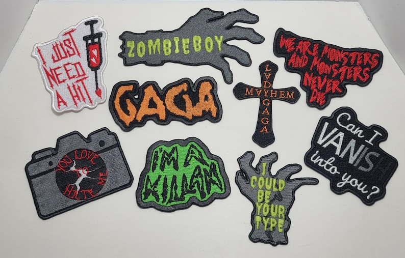 Puede incluir: Una colecci&oacute;n de parches bordados con varios dise&ntilde;os y texto. Los parches incluyen frases como "I just need a hit", "Zombieboy" y "Gaga". Otros parches presentan una c&aacute;mara, una cruz y una mano con texto. La paleta de colores incluye negro, gris, rojo, naranja y verde.
