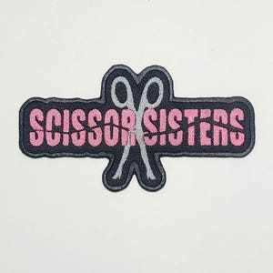 May include: Embroidered patch featuring the text "SCISSOR SISTERS" in pink, set against a black background. The design includes a pair of crossed scissors in the center, with a white outline. The patch has a black border.