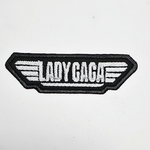 May include: A black and white embroidered patch featuring the text "LADY GAGA" in a stylized font. The patch has a rectangular shape with wing-like designs on each side, and a black border.