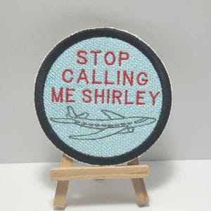 May include: A round blue and black embroidered patch with the text "STOP CALLING ME SHIRLEY" in red thread. A small airplane is embroidered in black thread in the center of the patch.