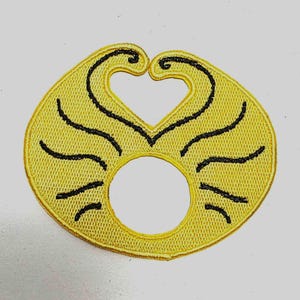 Puede incluir: Un parche bordado amarillo con forma circular y un recorte en forma de corazón en la parte superior. Líneas negras curvas irradian desde el centro, creando un diseño decorativo. El parche tiene una apariencia texturizada y es probablemente para ropa o accesorios.