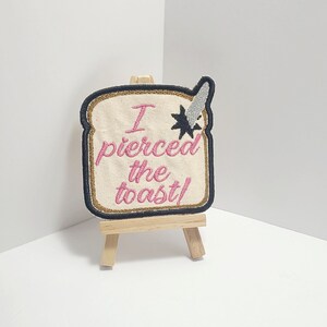 May include: A beige embroidered patch shaped like a slice of bread with the text "I pierced the toast!" in pink thread. The patch is displayed on a small wooden easel.