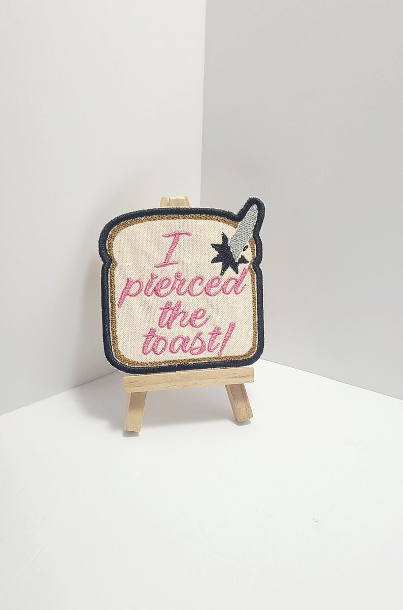 May include: A beige embroidered patch shaped like a slice of bread with the text "I pierced the toast!" in pink thread. The patch is displayed on a small wooden easel.