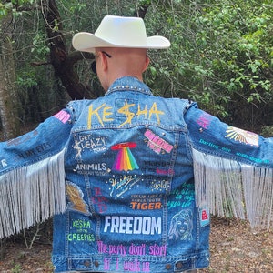 Puede incluir: Una chaqueta vaquera azul con flecos en las mangas y una variedad de parches y bordados coloridos. La chaqueta tiene el texto "Kesha" escrito en dorado en la espalda, junto con otras frases como "Get Leazy" y "Freedom".
