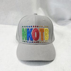 May include: A grey baseball cap featuring the text NKOTB in glittery, multi-colored letters. The letters are green, blue, red, orange, and yellow. The text is bordered above and below by a row of small, colorful, reflective rhinestones.