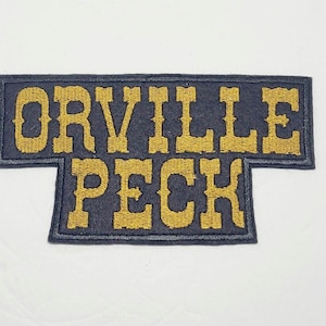 May include: A black and gold embroidered patch with the text "ORVILLE PECK" in a vintage style font.