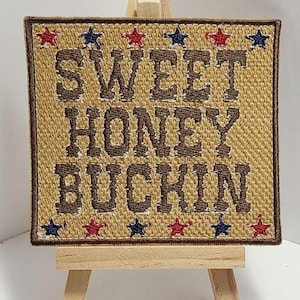 Peut inclure: Un patch brodé marron avec le texte "Sweet Honey Buckin'" en lettres marron. Le patch est entouré d'étoiles rouges et bleues.