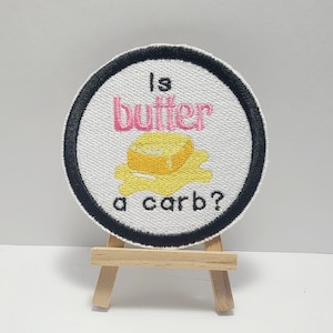 May include: A white round patch with a black border. The patch has pink text that says "Is butter a carb?" with a yellow stick of butter graphic.