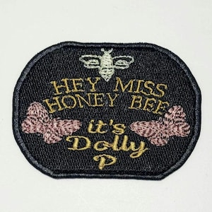 May include: Black embroidered patch with the text "HEY MISS HONEY BEE it's Dolly P" in gold thread. The patch features a bee and two pink and brown floral designs. The patch has a black border.