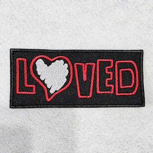 May include: A rectangular black embroidered patch featuring the word LOVED in red outline lettering. The letter O is replaced by a white heart shape with a red outline. The patch has a black border and is set against a light gray background.