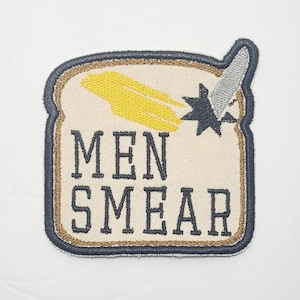 May include: A beige embroidered patch shaped like a slice of bread with a yellow smear of butter and a black and gray knife sticking out of the top. The text "MEN SMEAR" is embroidered in black on the patch.