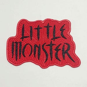May include: A red embroidered patch with the words "LITTLE MONSTER" in black, stylized lettering. The patch has a textured appearance and a unique shape, suitable for clothing or accessories.
