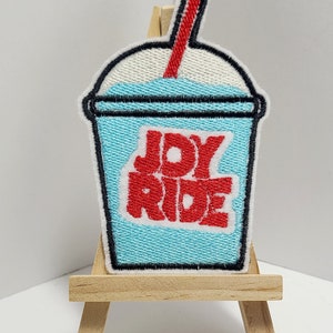 以下が含まれることがあります： 赤いストローと赤い文字「JOY RIDE」が書かれた、青と白の刺繍パッチの、スラリーカップ