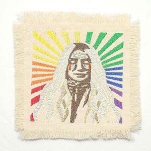 May include: A white fabric patch with a rainbow background and a stylized image of a person with long white hair and a rainbow-colored eye. The person is wearing a brown shirt and has a crown on their head.