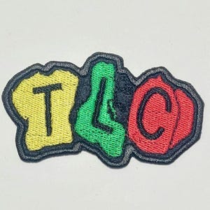 May include: Embroidered patch with the letters "TLC" in yellow, green, and red. The letters are outlined in black and have a textured appearance. The patch is designed to be sewn or ironed onto fabric.