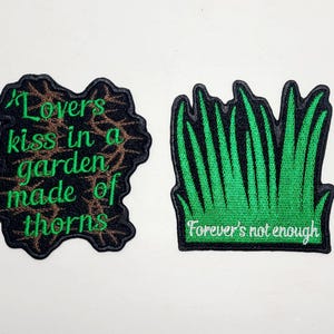 May include: Two embroidered patches. One patch features the text "Lovers kiss in a garden made of thorns" in green thread on a black background with brown accents. The other patch has green grass with the text "Forever's not enough".