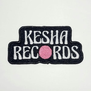 以下が含まれることがあります： 白字で「KESHA RECORDS」と書かれた刺繍パッチ。黒い縁取りがあり、テキストの中央にはピンク色の円があります。このパッチは黒い縁取りがあり、衣類やアクセサリー用です。